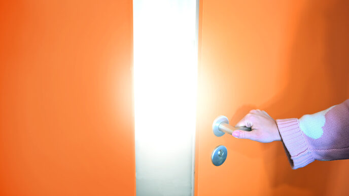 Eine Hand greift die Klinke einer etwas geöffneten orangefarbenen Tür, die den Blick auf ein helles Licht freigibt.