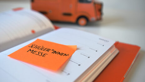Foto von einem orangen Post-It mit dem Wort "Schüler*innen-Messe" auf einem Kalender klebend. Im Hintergrund steht eine Stiftebox auf dem Tisch.