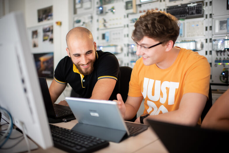 Foto von zwei Studierenden, die nebeneinander im Labor für Energieautomation und Netzführung an Arbeitsplätzen mit Computern sitzen. Sie tauschen sich aus.