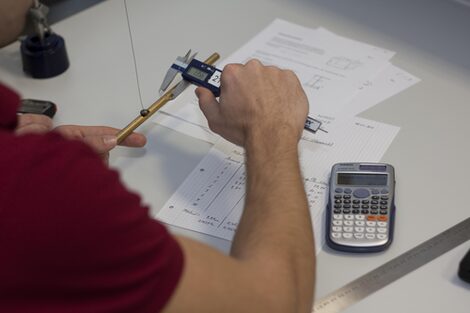 Foto einer Person, die mit einem Messschieber den Durchmesser eines Rohres bestimmt. Auf dem Tisch liegen Zettel mit Aufzeichnungen, ein Taschenrechner und ein Lineal.