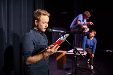 Foto eines Schauspielers im Theaterlabor auf der Bühne am Mikrofon, er liest aus einem Buch vor. Dahinter zwei weitere Personen, die auf Tischen sitzen