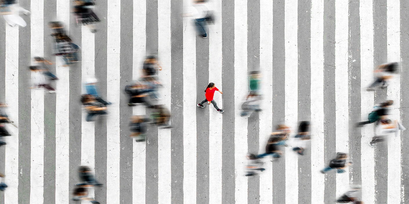 Blick von oben auf einen großen Zebrastreifen auf einer Straßen. Viele Menschen laufen drüber. Sie sind verschwommen dargestellt. Eine Person in einem roten Pullover ist mittig scharf im Fokus.