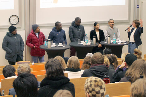 Seven people at high tables in front of well-filled rows of seats in the lecture hall look at each other. The person on the far right speaks into a microphone and gesticulates.