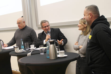 Four people at high tables, the second person from the left speaks into a microphone and gestures towards the people on the right.