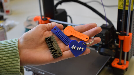 In einer Handfläche liegen vier verschieden gestaltete Schlüsselanhänger, im Hinterrund ist ein 3D-Drucker zu sehen.