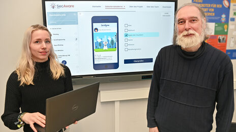 Zwei Personen stehen vor einem großen Bildschirm, auf dem die digitale Lernplattform "SecAware".nrw zu sehen ist.