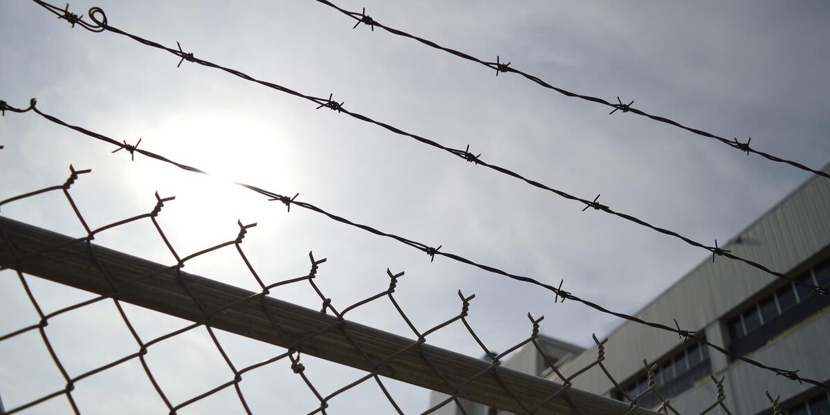 A high fence with barbed wire. A prison building can be seen in the background.
