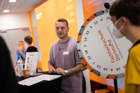Next to a wheel of fortune with Fachhochschule Dortmund written on it, a young male person stands talking to some prospective students.