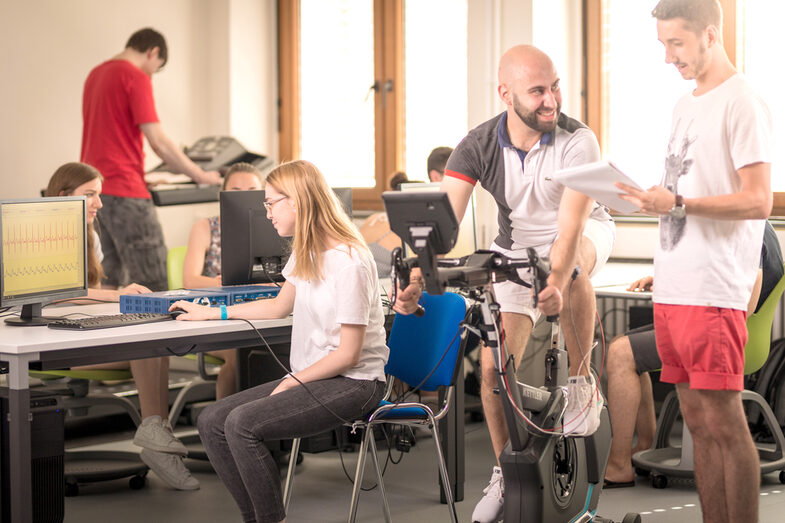 On the right in the photo, a man on a bicycle ergometer and another man next to him taking down values. On the left in the photo, other people at computers.