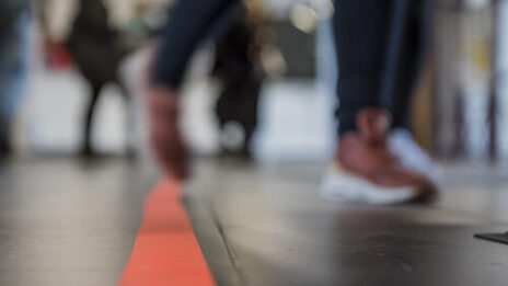 Foto einer orangene Linie auf dem Boden sowie Füße mehrerer Menschen, die über den Flur laufen. __ <br>Orange line on the floor and feet of several people walking down the hall.