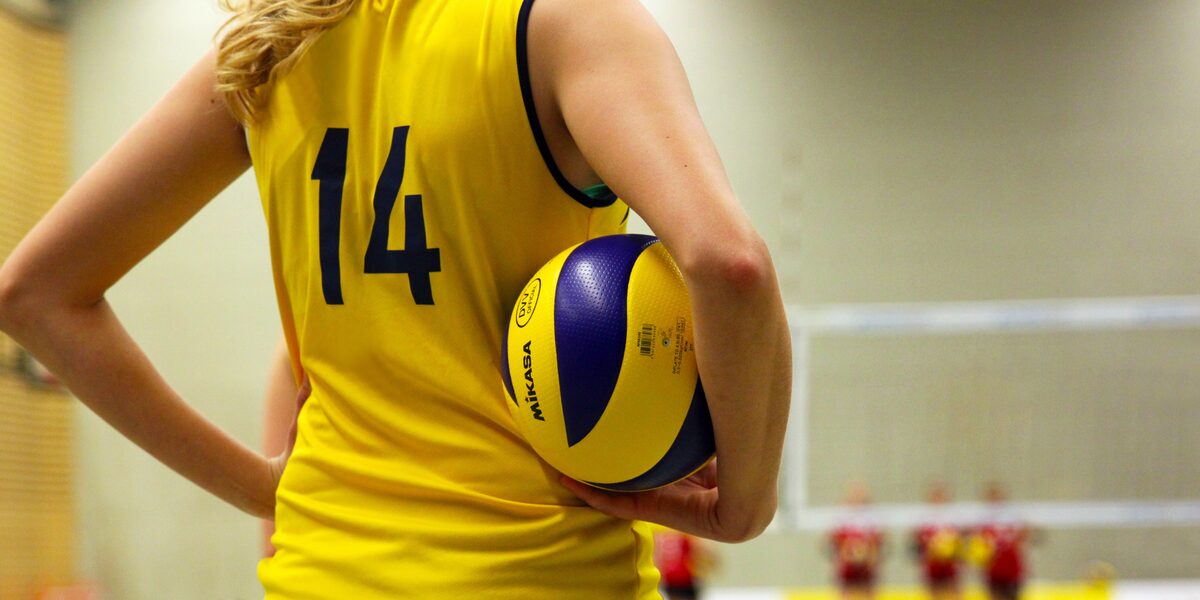 Eine weiblich gelesene Person steht in einer Sporthalle auf einem Volleyballfeld und hält einen Ball unter dem Arm.