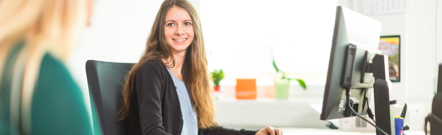 Photo of a young woman at a workplace with a computer. She looks at another woman at a desk who is taking notes - she is only partially recognizable.