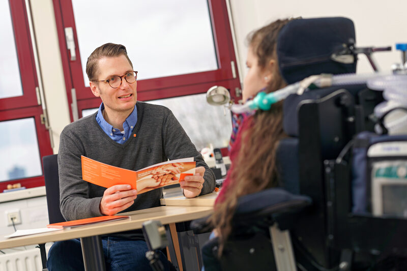 Foto einer Frau in einem Elektrorollstuhl, die mit einem Mann ein Beratungsgespräch über barrierefreies studieren führt. Der Mann hält Broschüren in der Hand.