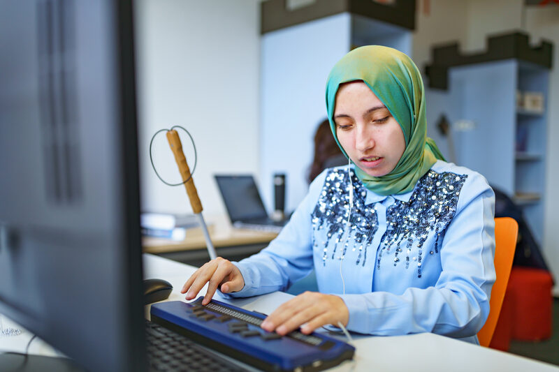 Foto einer Studentin mit Sehbehinderung, sie arbeitet an einem Computer mit Braillezeile. Neben ihr steht ein Blindenstock.