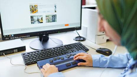 Foto der Hände einer Studentin mit Sehbehinderung, die auf der Braillezeile vor einem Computerbildschirm abgelegt sind.