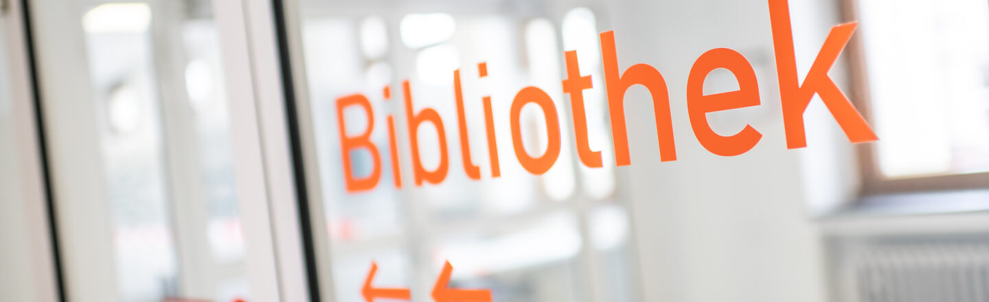 Foto von orangener Folienschrift "Bibliothek" und Pfeilen, auf einer Glastür.