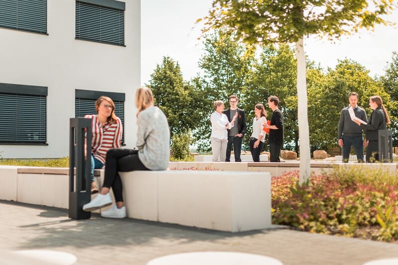 Foto von mehreren Personen, die auf dem Campus stehen und sitzen. Darunter Doktorandeninnen und Doktoranden sowie Promovierende.