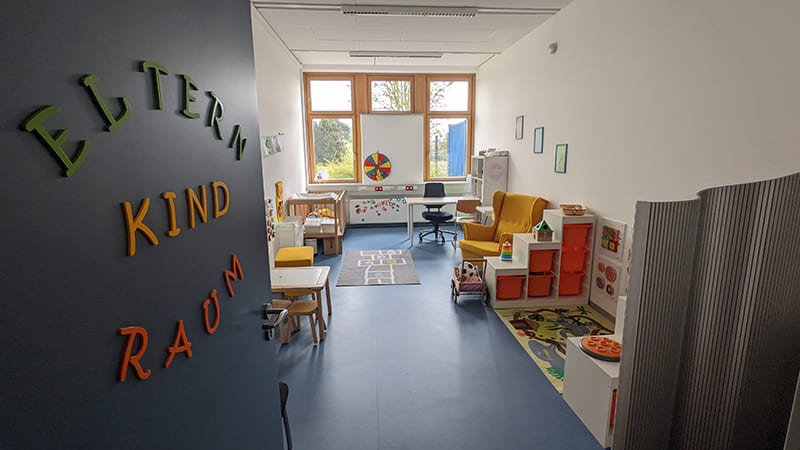Foto von einem Eltern-Kind-Raum, die Tür ist geöffnet und man schaut in einen länglichen Raum, links steht ein Tisch und weitere Möbel, rechts sind Regale, ein Spielteppich und ein gelber Sessel sowie hinten ein Schreibtisch für Erwachsene.