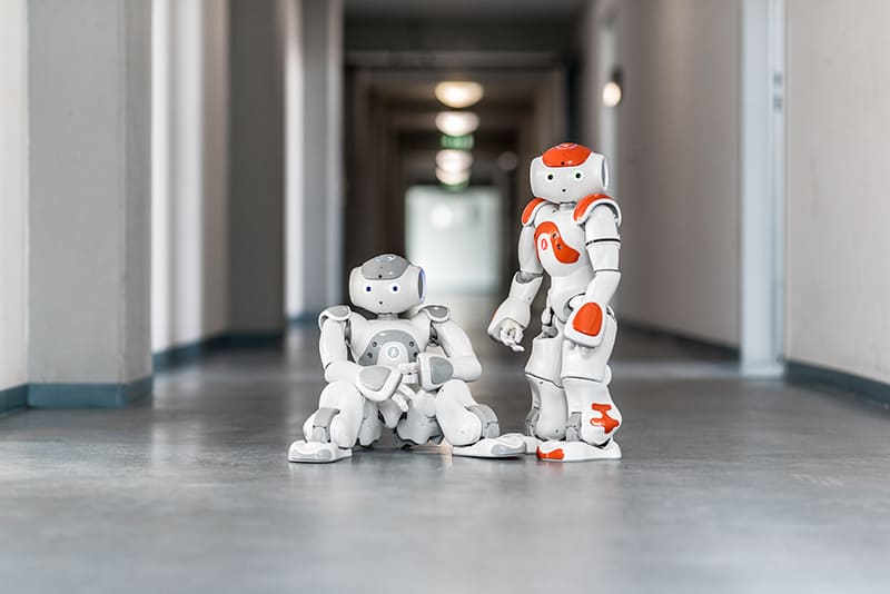 Foto von zwei NAO Roboter auf einem Flur. Der rechte sitzt, der Linke steht.