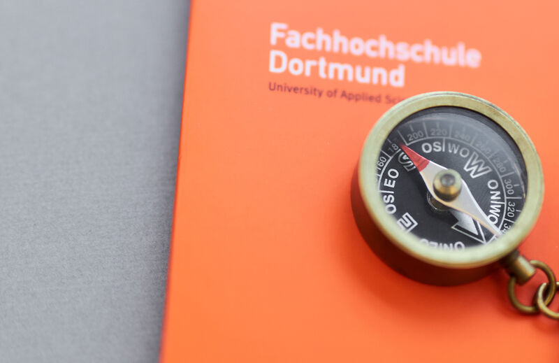 Foto eines kleinen Kompasses mit Kette daran, der mit einem Bleistift auf einer orangefarbenen Mappe der Fachhochschule Dortmund liegt.