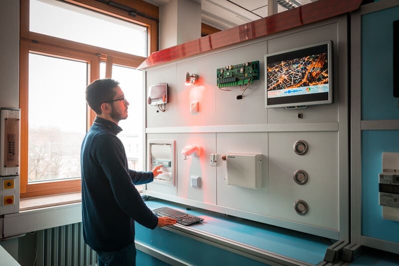 Foto eines Aufbaus eines Smart Buildings. Die Wand hat oben ein Dach. Lichter und Monitor sind aktiviert. Ein Studierender steht davor und bedient eine Lampe mit einem Schalter.