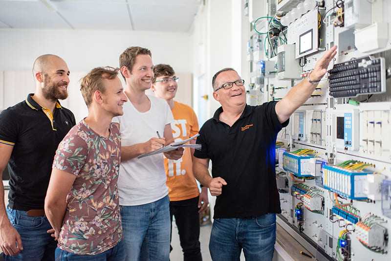 Foto eines Professors, der vor einer Versuchswand der Elektrotechnik steht und auf ein Gerät zeigt. Neben ihm stehen vier lächelnde Studenten, einer von ihnen macht sich Notizen.