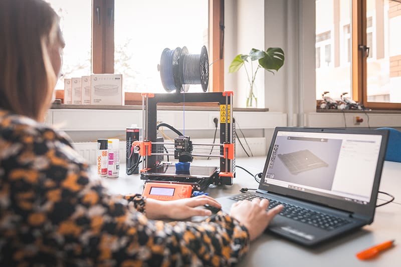 Foto einer Studentin, die auf einen 3D-Drucker schaut, der gerade etwas druckt. Nebenbei tippt sie am Laptop und bedient den Drucker.