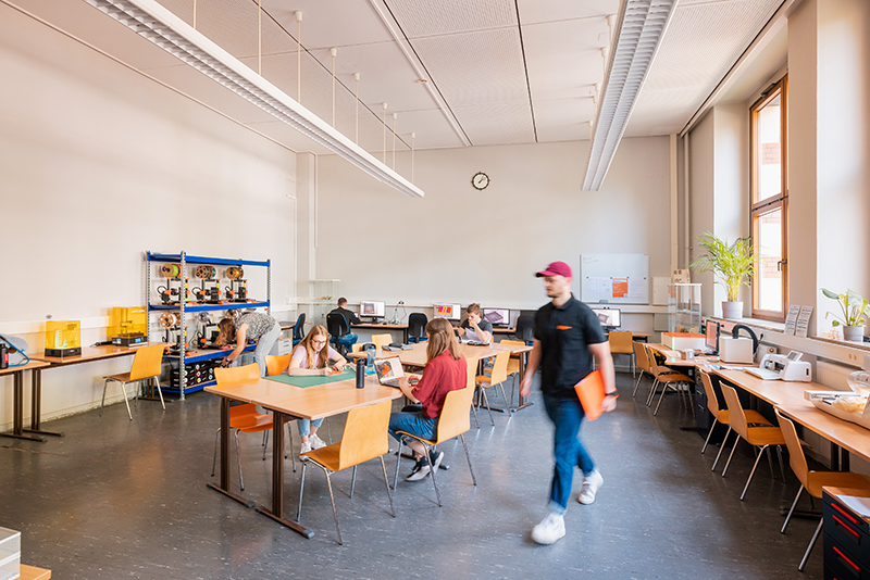 Raumaufnahme von einem Werkraum in dem verschiedene Arbeitsplätze mit verschiedenster Technik ist, z.B. 3D-Drucker, Computer, Laser, Plotter, Nähplatz. Im Raum arbeiten Studierende an verschiedenen Stationen.