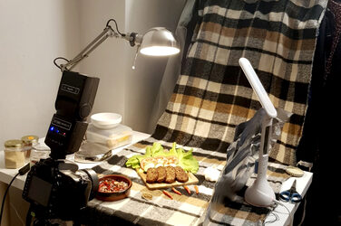 Eine Kamera und Lampen sind auf einen Tisch gerichtet, auf dem Lebensmittel zom Fotografieren drapiert sind.