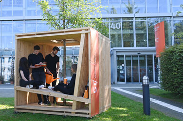 People sit in a large wooden cube in front of the Fachhochschule Dortmund architecture building. The cubes are part of the anniversary campaign.