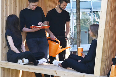 People sit in a large wooden cube in front of the Fachhochschule Dortmund architecture building. The cubes are part of the anniversary campaign.