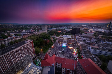 The aerial photograph shows Dortmund's Friedensplatz, where numerous people are standing in front of a stage. The sky has a color gradient from bluish to yellowish.
