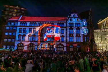 A streetcar with two eyes is projected onto the façade of a building. Several people are watching the scene.