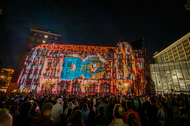The letters DO are projected onto the façade of a building and several people follow the scene.