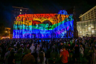 A building façade is illuminated in rainbow colors, the letters DO can be seen in the middle. Several people are watching the scene.