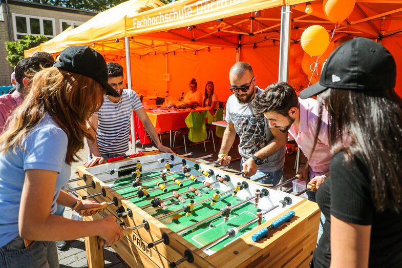 Several people are standing around a table football table at a summer party. Four people are playing table football against each other, two people are watching them.