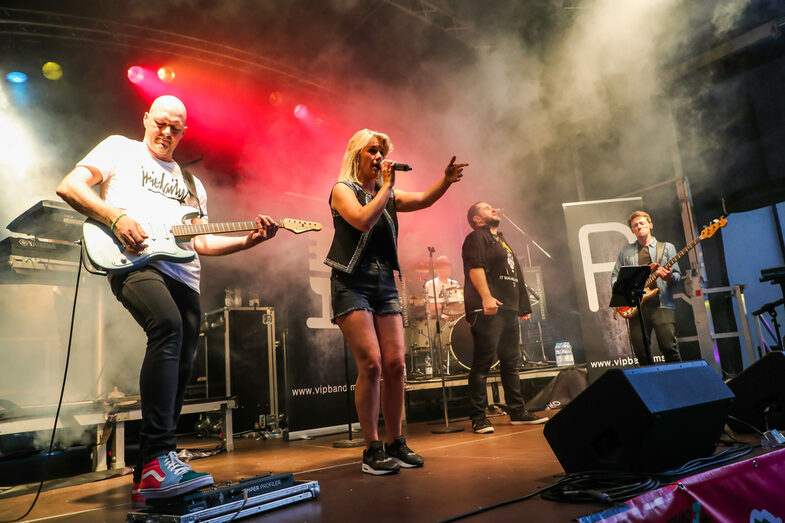 A music band is playing on a stage. Two men are playing guitar, a woman and a man are singing into a microphone. At the back of the stage, a man is sitting at a drum kit.