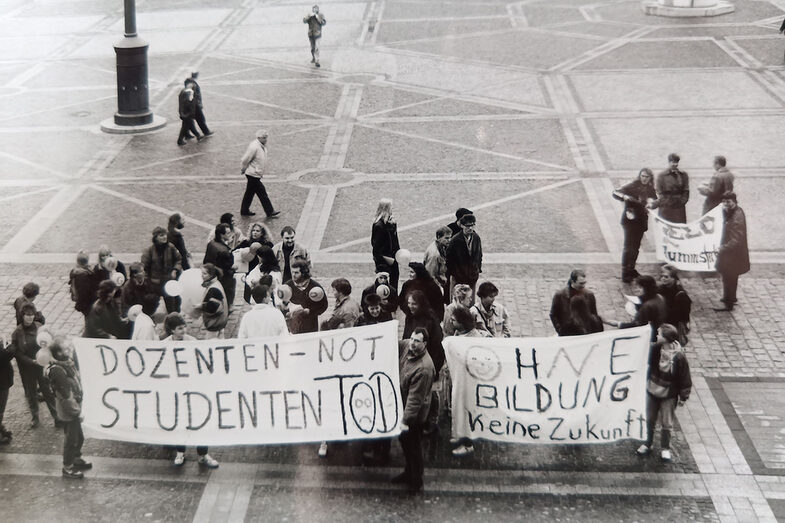 Eine kleinere Gruppe Menschen steht auf einem Platz und hält Transparente. Auf einem steht "Dozenten-Not = Studenten Tod" auf einem zweiten "Ohne Bildung keine Zukunft".