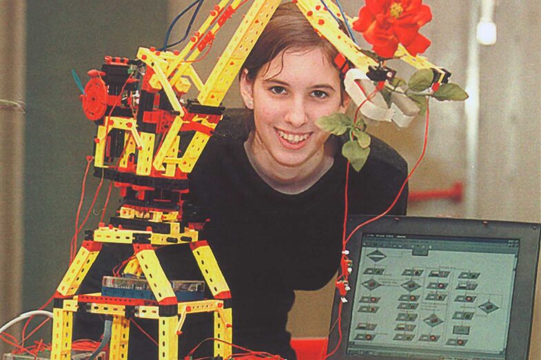 A young woman next to a kind of toy crane with a gripper arm. Next to it is a laptop. The robotic arm is holding a rose.