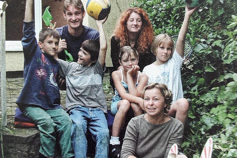 Four children and three adults are sitting on an outside staircase in front of a building. The children are holding up balls.