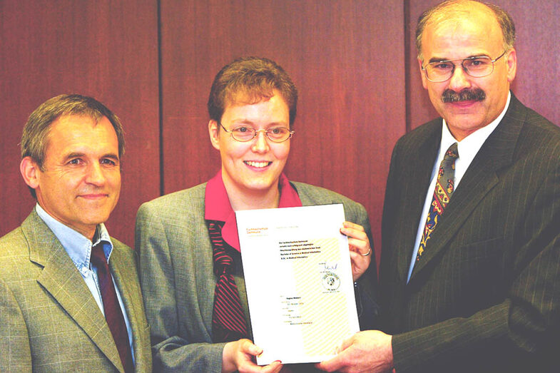 Three people stand next to each other and look towards the camera. The woman in the middle holds up a certificate.