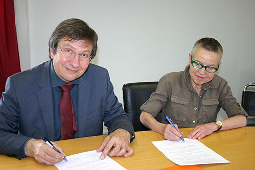 Two people, a man and a woman, sit at a desk and sign papers.