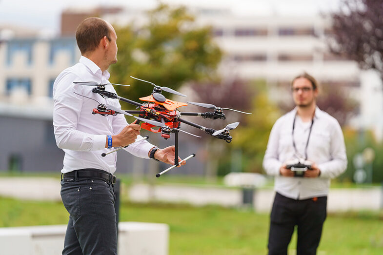 One man holds an orange drone in his hand, a second man holds the controls for the drone. They stand opposite each other.