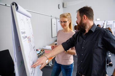 Foto von einer Frau und einem Mann, die bei einem Seminar der Personalentwicklung vor einem Flipchart stehen, der Mann zeigt etwas darauf.__Photo of a woman and a man standing in front of a flipchart at a staff development seminar, the man is pointing something on it.