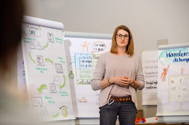 Photo of a woman standing in front of several flipcharts with colorful drawings and labels and telling something __Photo of a woman standing in front of several flipcharts with colorful drawings and labels and telling something.