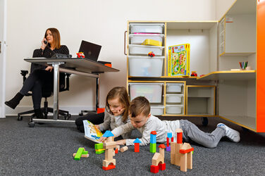 Photo of two younger children playing with building blocks on the carpeted floor of an office. In the background, their mother is sitting at a desk and talking on the phone.__Photo of two younger children playing with building blocks on the carpeted floor of an office. In the background, their mother is sitting at a desk and talking on the phone.