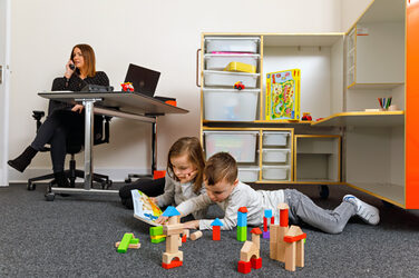 Foto von zwei jüngeren Kindern, die auf dem Teppichboden eines Büros mit Bauklötzen spielen. Im Hintergrund sitzt ihre Mutter an einem Schreibtisch und telefoniert.__Photo of two younger children playing with building blocks on the carpeted floor of an office. In the background, their mother is sitting at a desk and talking on the phone.