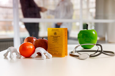 Foto eines Stilllebens aus verschiedenem Merchandise des Gesundheitsmanagements, wie zum Beispiel dem Antistressball. Ansonsten Impfpass, Stethoskop und Äpfel.