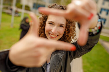Foto einer Studentin, die mit Zeigefingern und Daumen vor sich ein Quadrat formt und dadurch guckt.