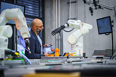 Foto von einem Labormitarbeiter, der am Transportband zwischen zwei Robotergreifarmen steht und eine Fernbedienung in der Hand hat.